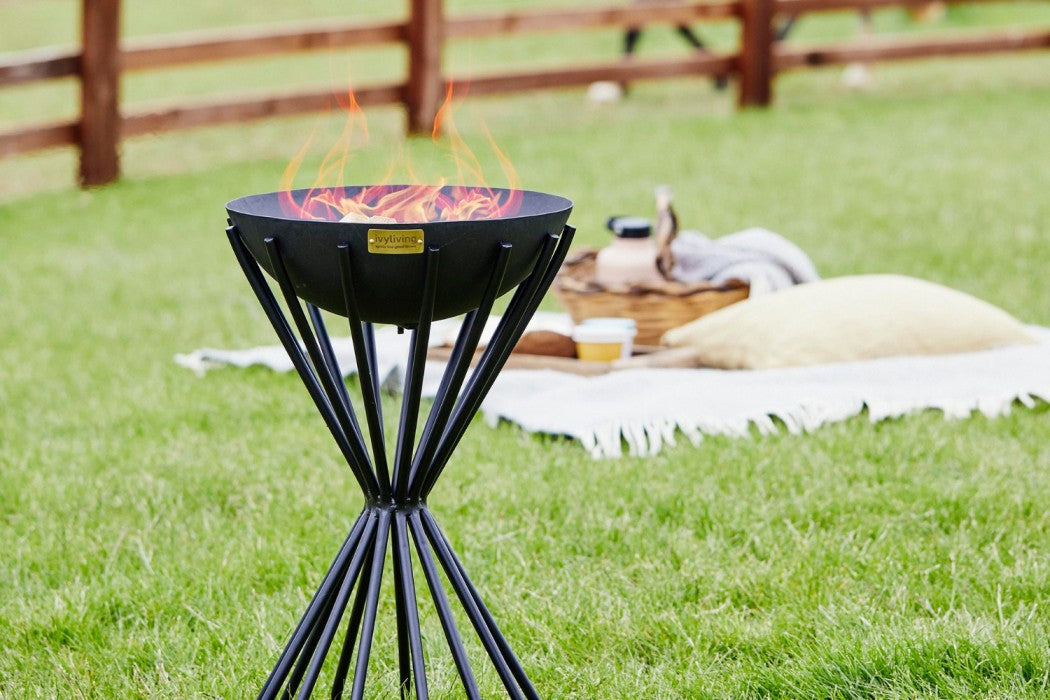 An Ivyline Tall Dakota  Firebowl on a grass lawn, in front of a picnic setting.