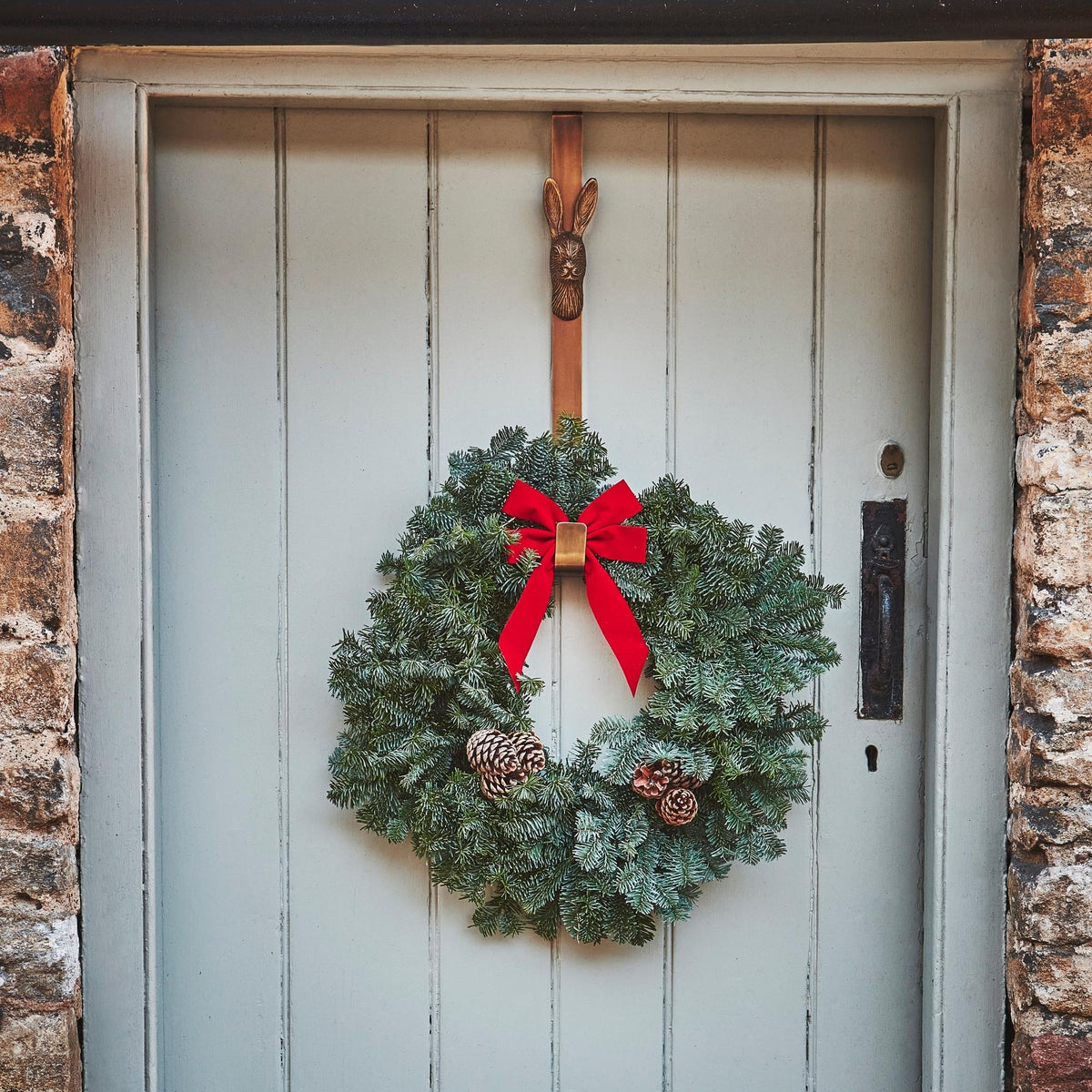 A brass wreath hanger with a festive Christmas wreath featuring green holly, red ribbons, and brown pinecones, hung on a gray door with a brick wall.